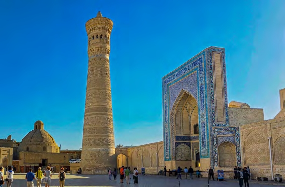 166 Tall brick minaret beside a blue-tiled, arched entrance in a historic Central Asian square, with tourists walking around.