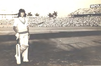 90 Cricketer in white with pads and bat standing on a cricket pitch as crowd watches from the stands.