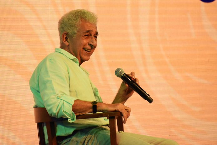 Older man in a light green shirt smiling while speaking into a handheld microphone on stage.