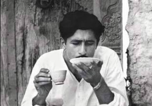 Black-and-white photo of a man in a white shirt sipping from a small cup, holding a saucer in his other hand.