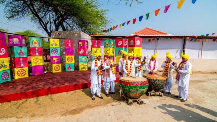 Group of men in white outfits and yellow turbans playing drums and flutes at a colorful festival backdrop.