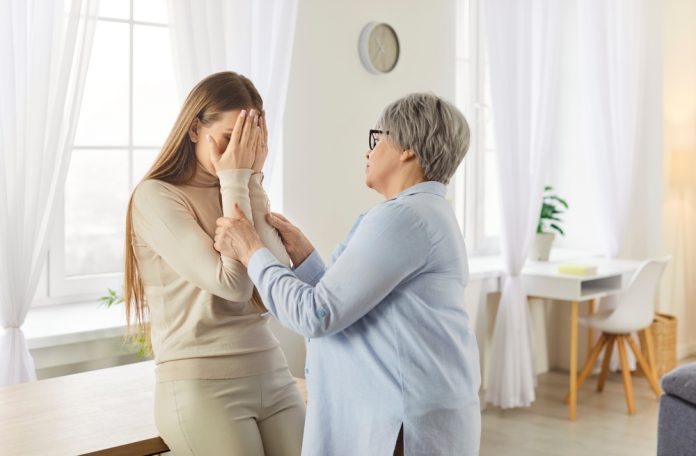 Older woman in a light blue blouse comforts a younger woman who covers her face with her hands in a bright home setting.