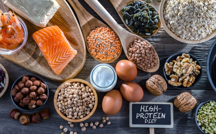 Food,Products,Rich,In,Protein.,High-protein,Diet Assorted high-protein foods on a wooden table (salmon, eggs, nuts, seeds, yogurt, oats) with a 'High-Protein Diet' sign.