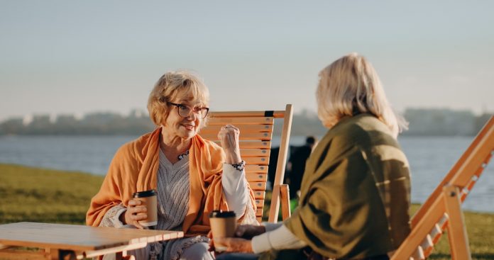 Two,Senior,Women,Friends,Sitting,In,Lounge,Chairs,,Holding,Coffee Two older women chat at a lakeside, each with a takeaway coffee, seated in wooden lounge chairs on a sunny day.