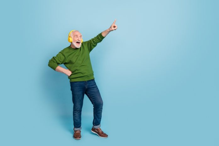 Full,Size,Portrait,Of,Positive,Carefree,Aged,Person,Look,Direct Older man in a green sweater and jeans wears yellow headphones, dancing and pointing upward against a blue backdrop.