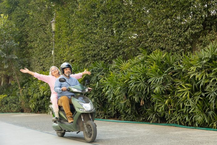 Older man wearing a helmet rides a green scooter while a cheerful woman sits behind him with arms outstretched on a tree-lined street.